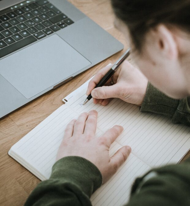 person at desk with laptop writing in notebook