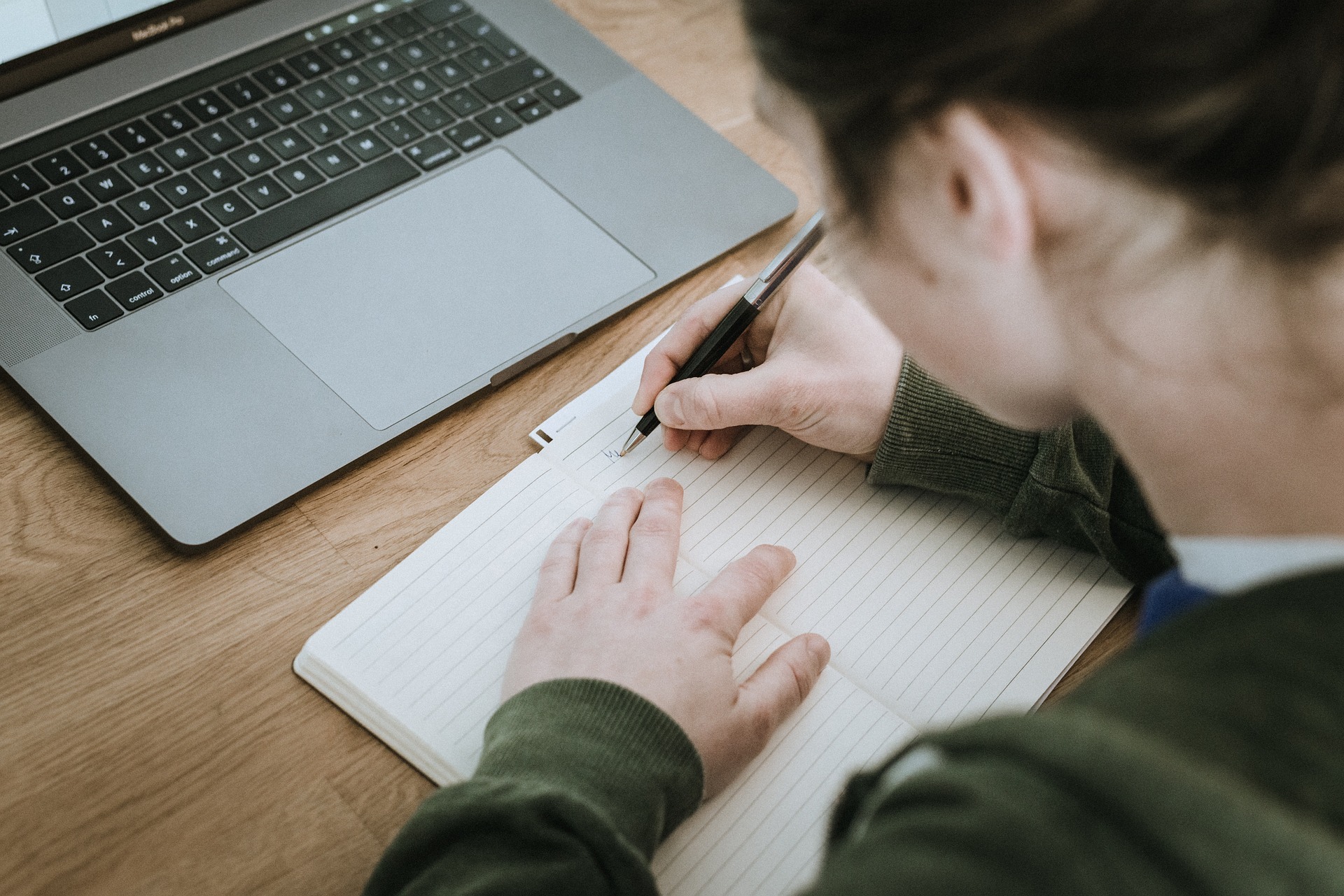 person at desk with laptop writing in notebook