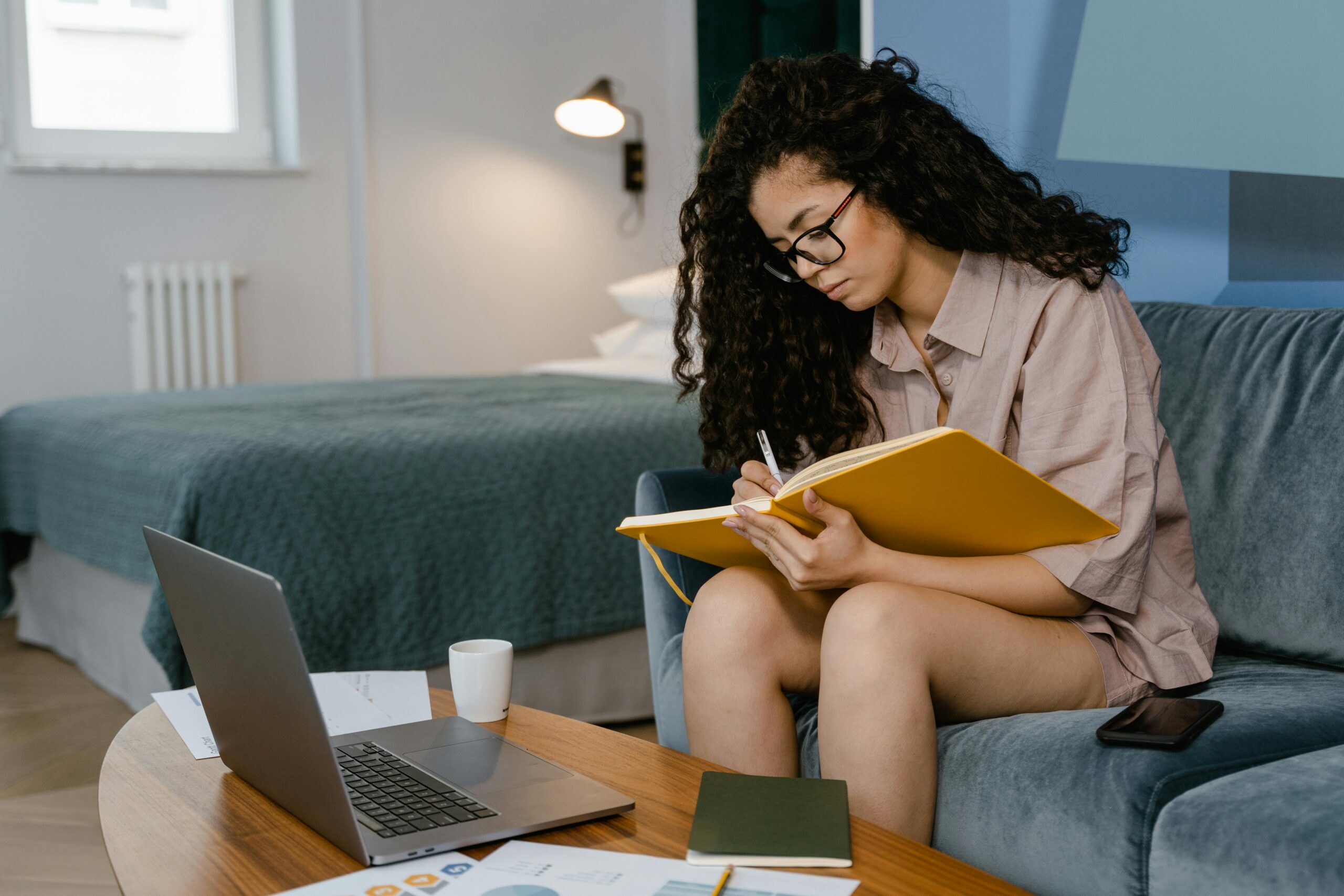 young woman with long, curly hair writing in notebook facing a desk with a laptop open on it