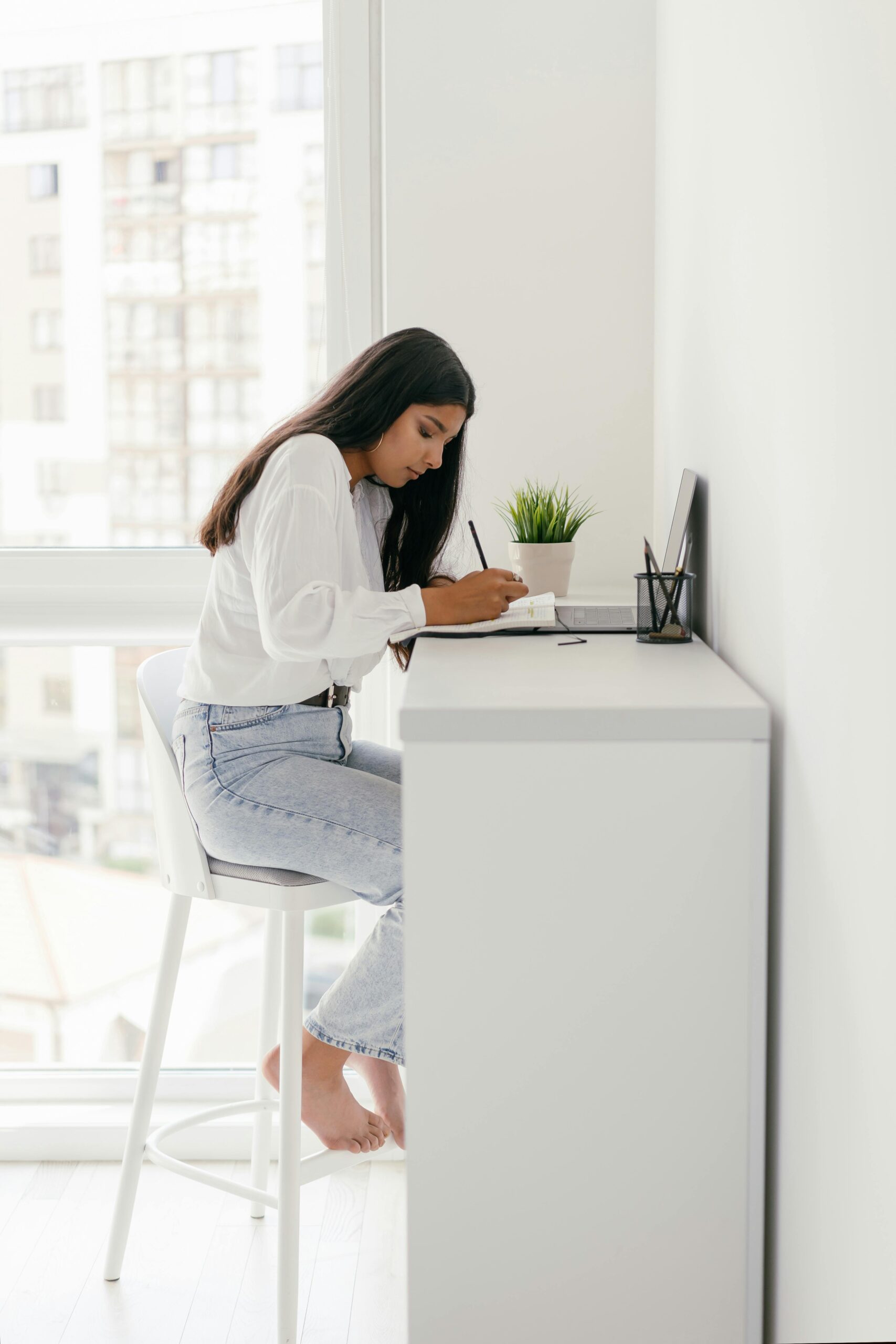 young woman sitting at high white desk writing in a notebook in front of a window