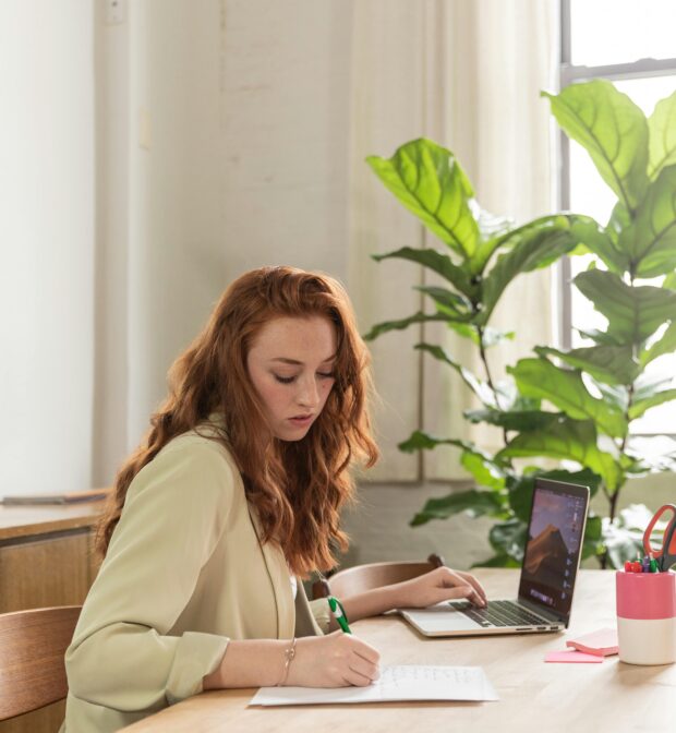 young woman writing in notebook and working at laptop at a desk with a plant on it