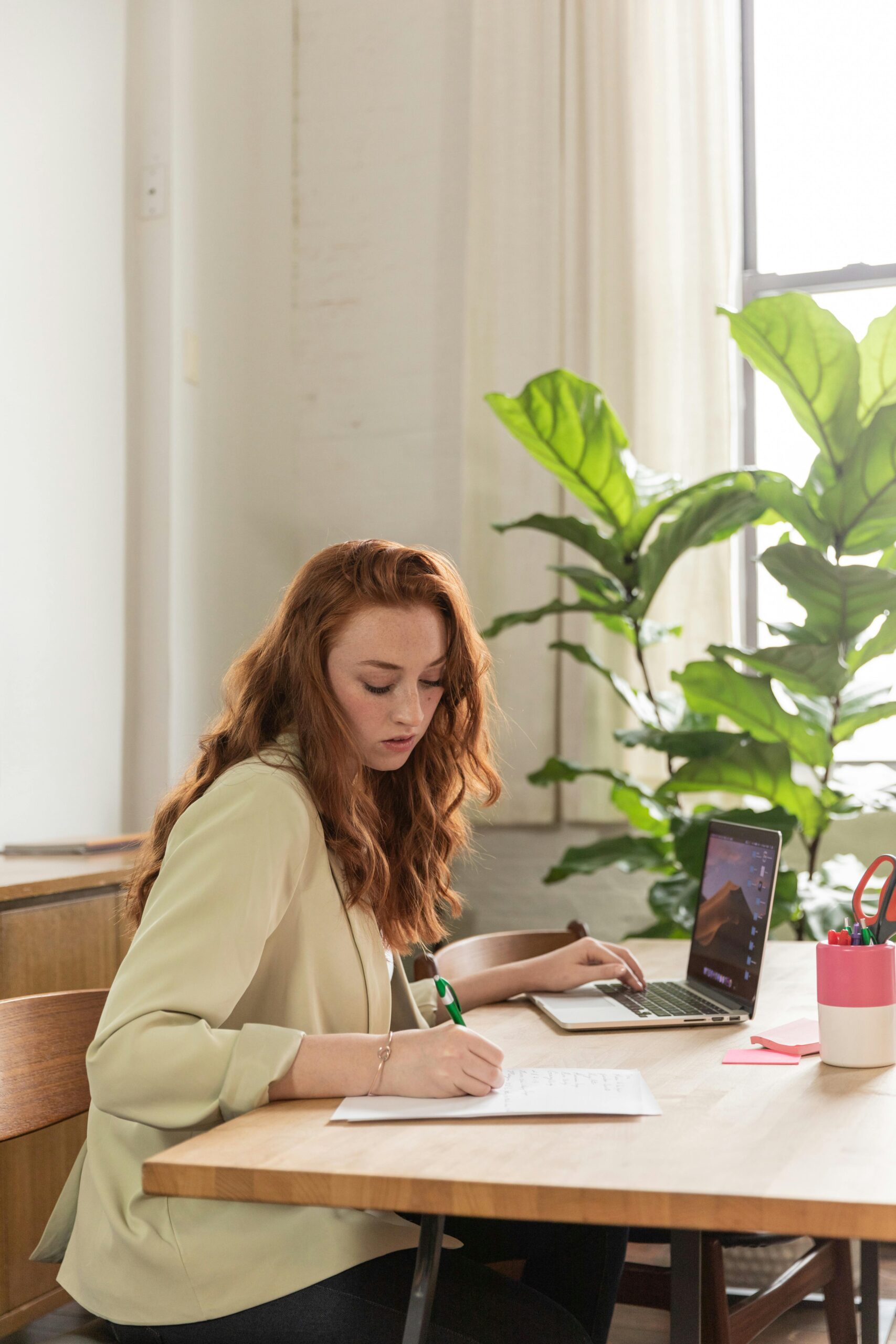 young woman writing in notebook and working at laptop at a desk with a plant on it