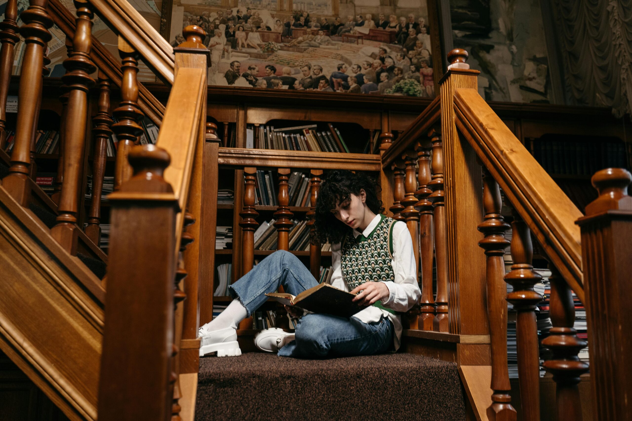 college student reading a book on the steps inside a library with books in the background