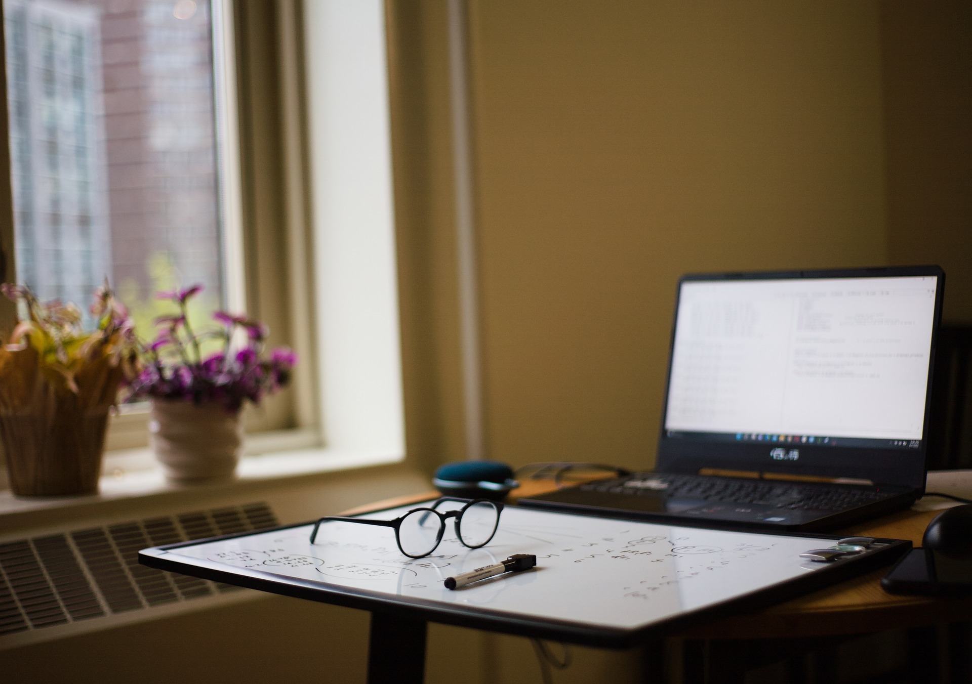 glasses, a whiteboard and a marker in front of an open laptop, next to a window