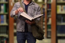 Person with a denim jacket and a shoulder bag reading a book in a library. Shelves filled with blurred colorful books are in the background.