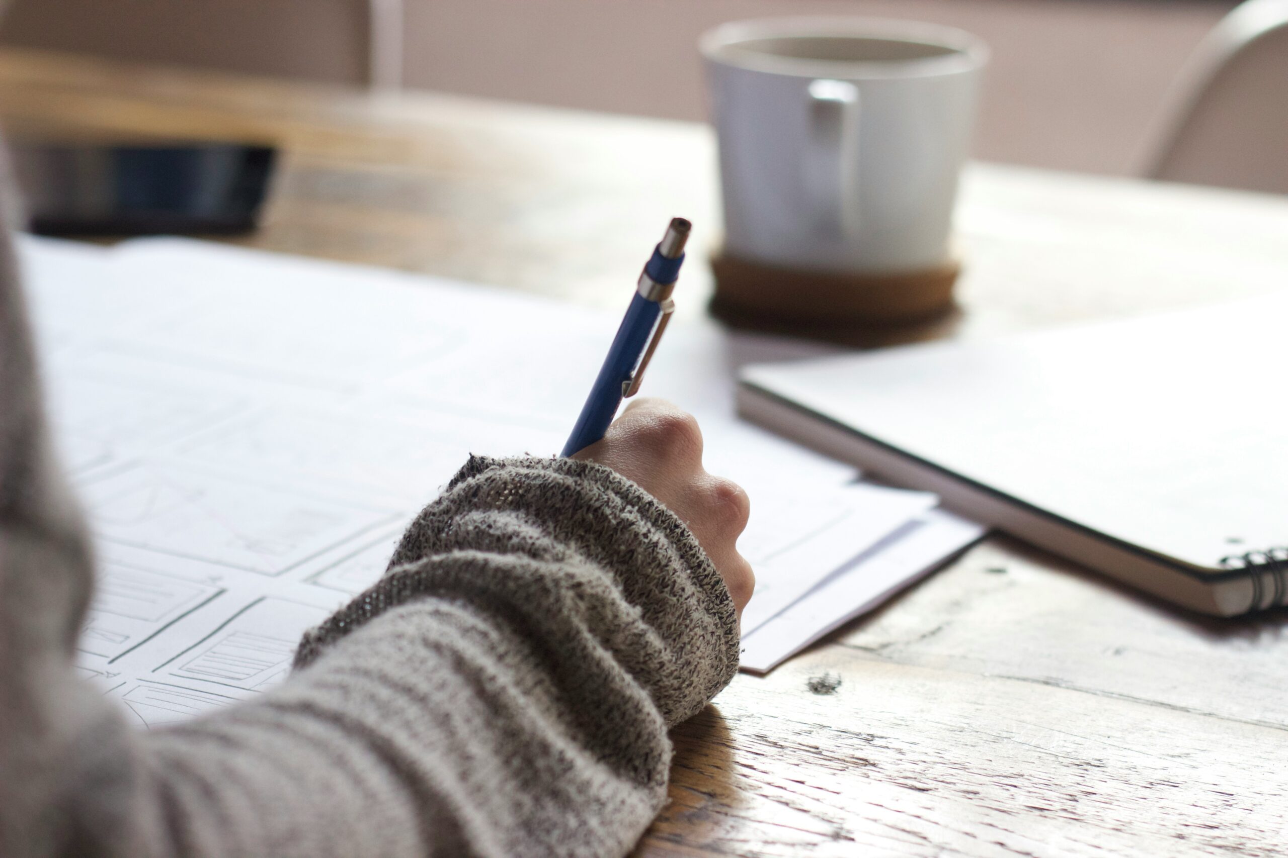 Close up of hand resting on wooden desk, writing with pen on paper with coffee cup blurred in background.