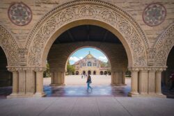 Ornate stone arch at Stanford University frames the colorful facade of Memorial Church. People walk in the sunlit courtyard, creating a serene atmosphere.