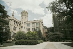 Historic university building with Gothic architecture, ivy-clad walls, and a central tower. Surrounded by lush greenery under a cloudy sky. Calm atmosphere.