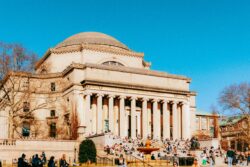 A grand neoclassical building with a large dome and columns stands against a clear blue sky. People sit on steps and grass, enjoying the sunny day.
