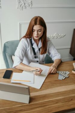 A focused doctor in a white coat sits at a desk, writing on a notepad. A stethoscope hangs around her neck. Nearby are a smartphone, tablet, and medicine packets.