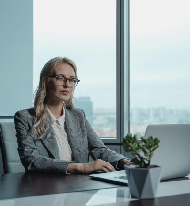 Woman in gray blazer working at laptop in office with skyline view in background