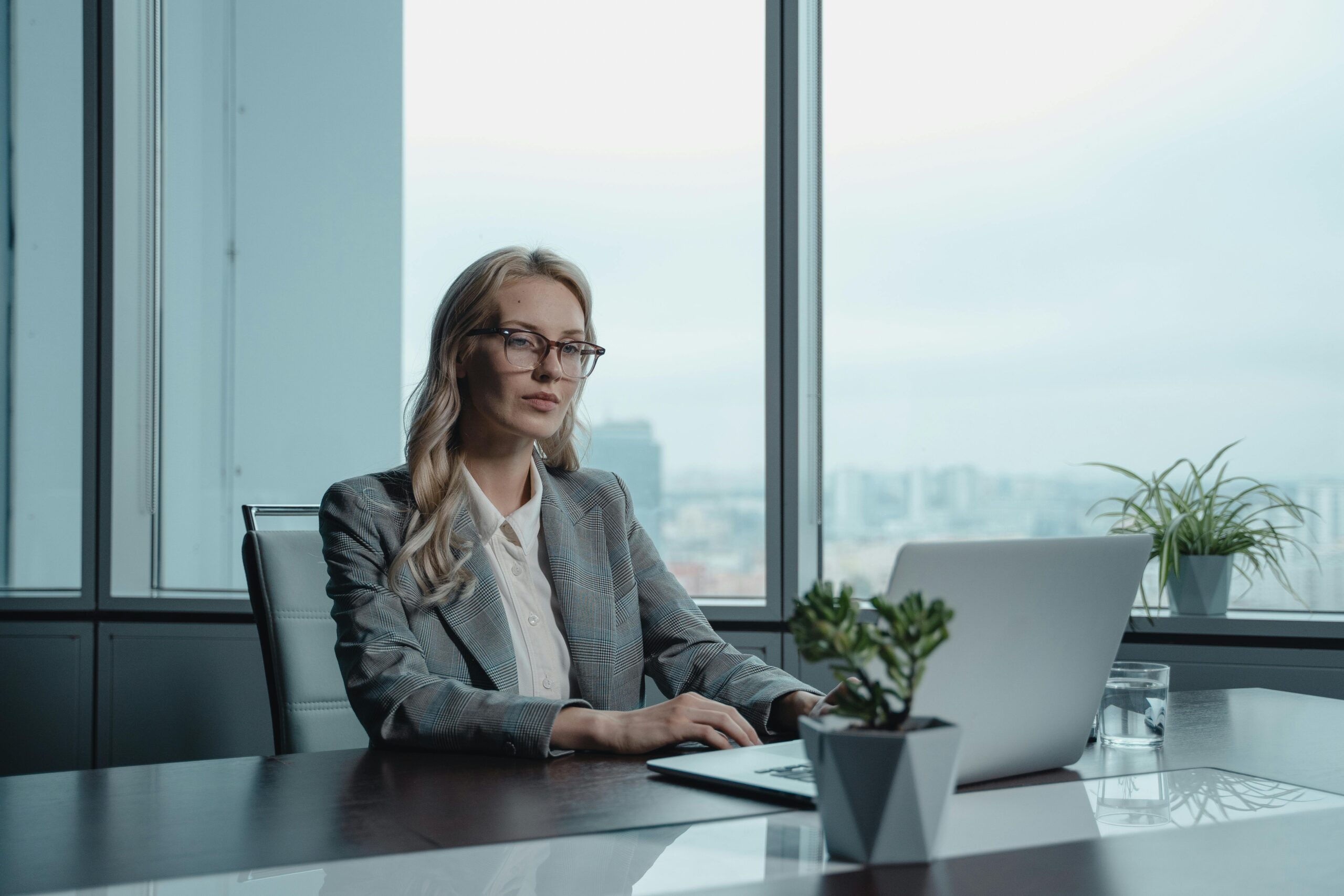 Woman in gray blazer working at laptop in office with skyline view in background