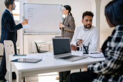 Two people standing by whiteboard with various countries listed, while two other people sit at a desk in discussion
