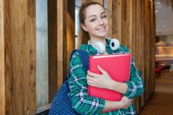 Smiling young woman with long hair, holding a pink notebook and wearing headphones around her neck. She's dressed in a green plaid shirt. Bright and cheerful mood.