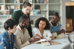 A group of five young adults sitting at a table in a library or study space, engaged in a focused discussion around an open book, with notebooks and shelves of books in the background.