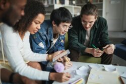 Group of young adults collaborating on a project at a table, surrounded by notes, paper, and a smartphone, with focused expressions as they write and discuss ideas in a cozy indoor setting.