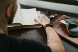 A young man with a leather bracelet on his right wrist sits at a table, reading a book. A watch and a violet flower are placed onside him.