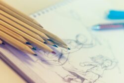 A group of color pencils lies on a sketchbook with faint drawings of people. The scene conveys creativity and focus, with a soft, artistic light.