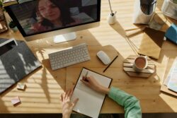 A student sitting at a study table, attending a virtual class on a Mac computer led by a female teacher. And writing notes in a notebook with a coffee cup nearby.
