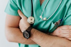 A young male doctor in green scrubs facing the camera, wearing a black watch on his left wrist.