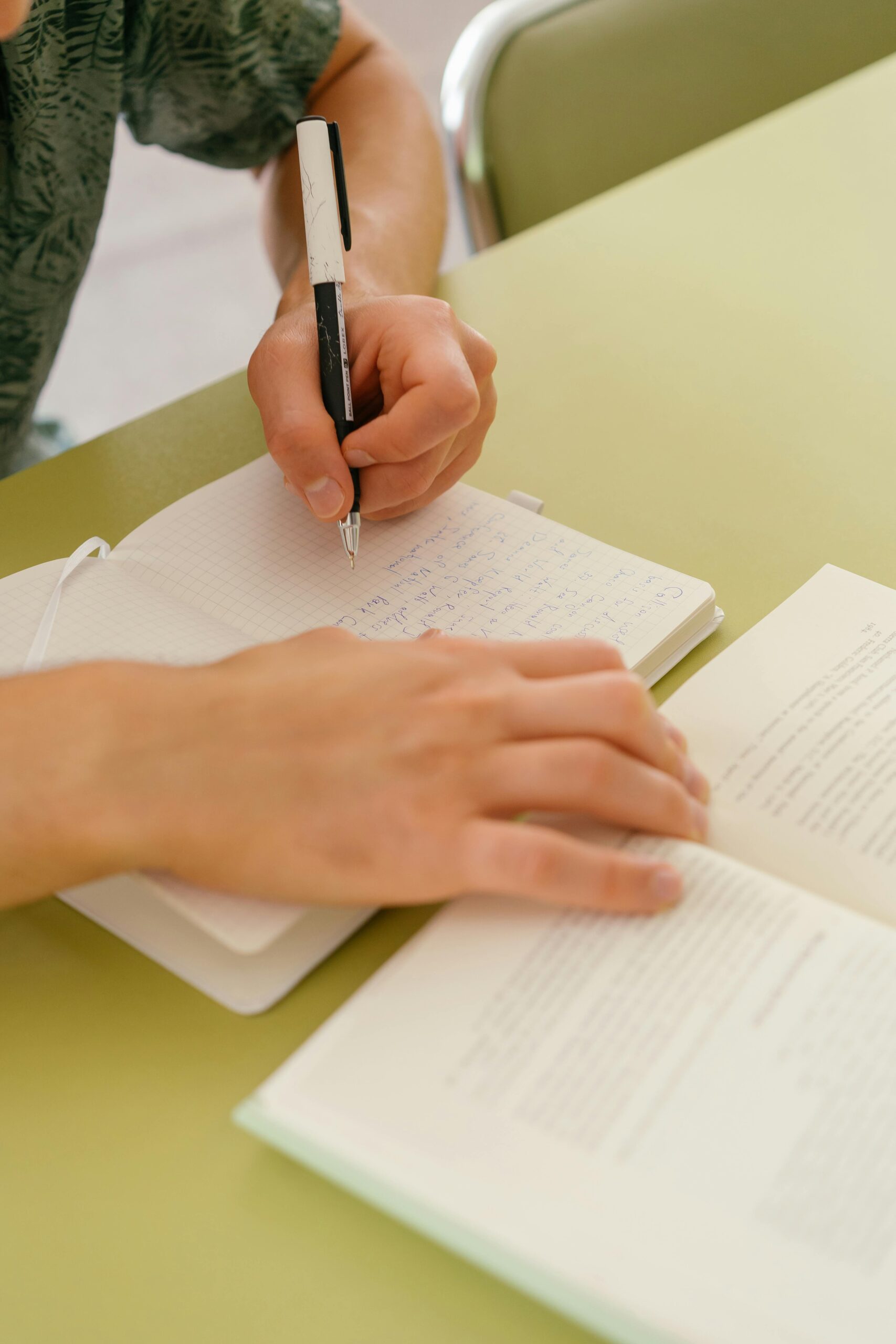 Close-up of a person writing in a notebook with a pen while referencing an open book, seated at a green desk, focusing on study or research work.