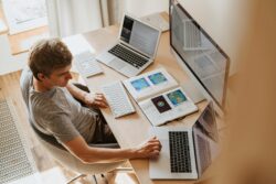 Young man studying geoscience at a desk with two laptops, a Mac PC, and an open notebook.