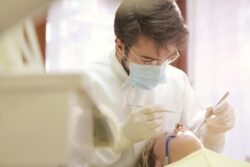 A young male dentist wearing a white mask examining a patient’s teeth in a dental clinic.