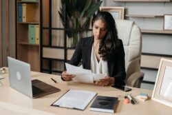 Woman in business attire sitting at desk with laptop and various papers, notebooks, and pens