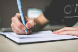 A hand holds a blue pen, writing in a notebook on a desk. The setting is calm and focused, suggesting concentration and creativity.