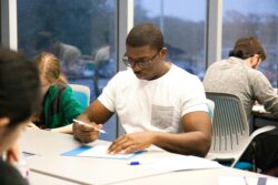 A focused student in glasses, wearing a white shirt, writes attentively at a classroom desk. Nearby, others are seated, highlighting a study atmosphere.