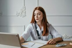 A focused woman in a doctor's coat works at a desk with a notebook, pen, and laptop. Blister packs of pills are nearby, suggesting a medical setting.
